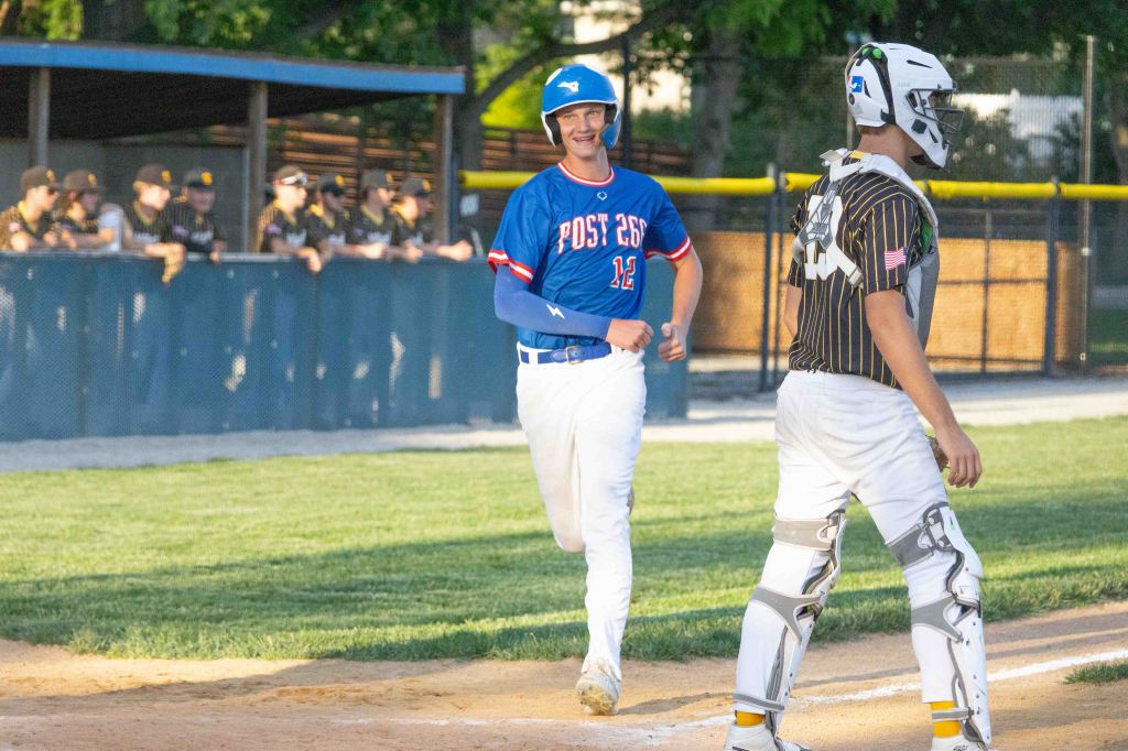 A baseball player in a blue jersey with the number 12 smiles as he approaches home plate, while a catcher in a black and white striped uniform watches. In the background, teammates in brown uniforms are observing from the dugout.