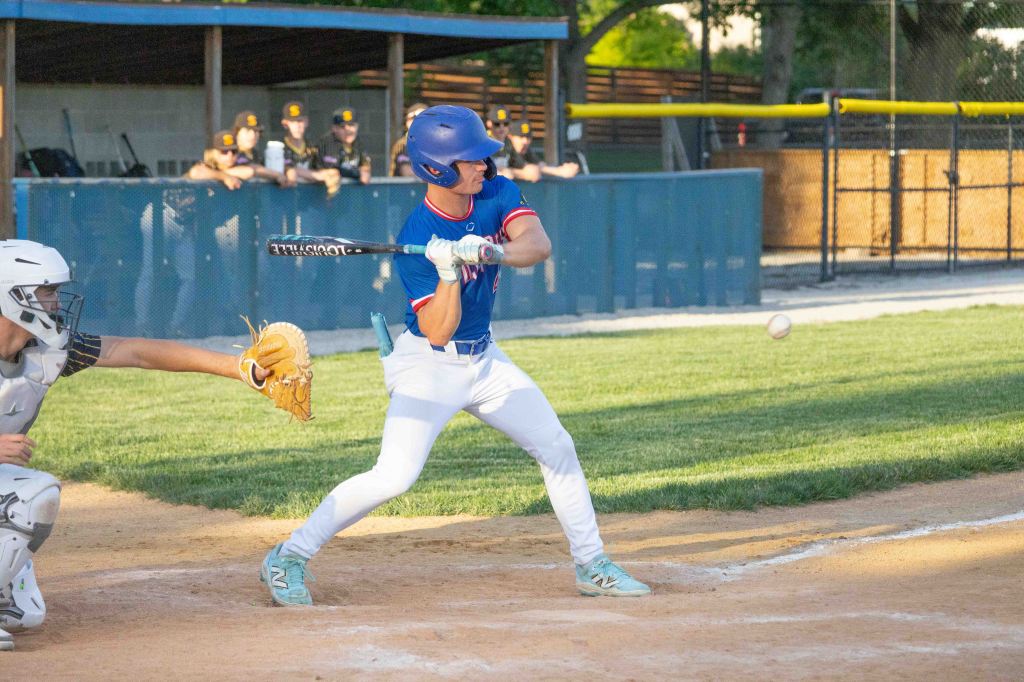 A young baseball player swings a bat during a game, with a catcher and teammates visible in the background.