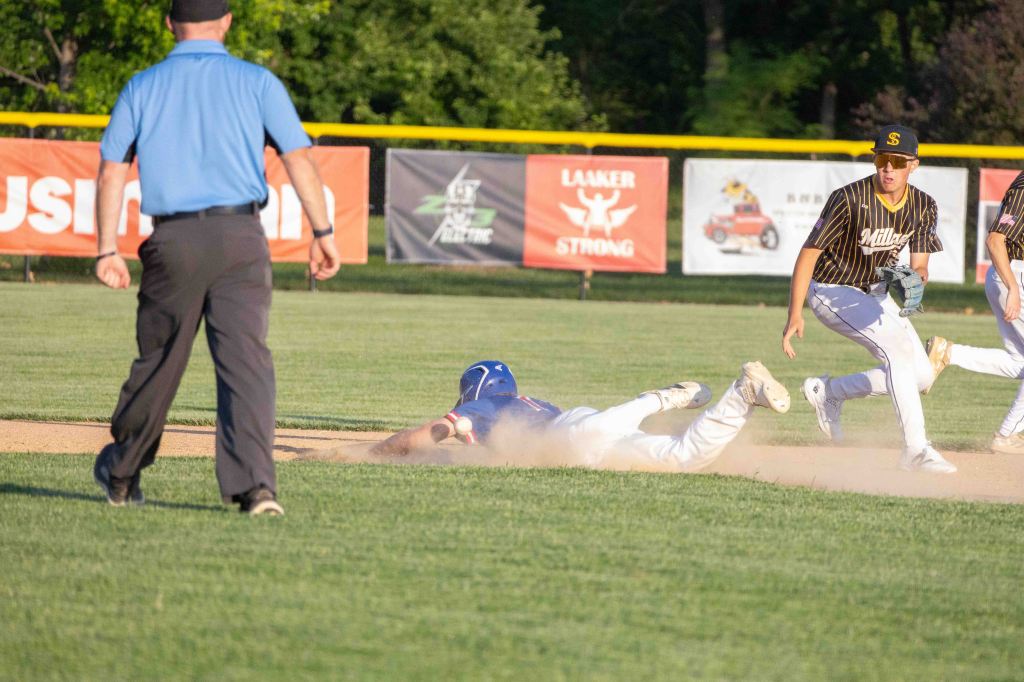 A baseball player diving to reach a base, kicking up dust, with an umpire observing the play and other players in the background.