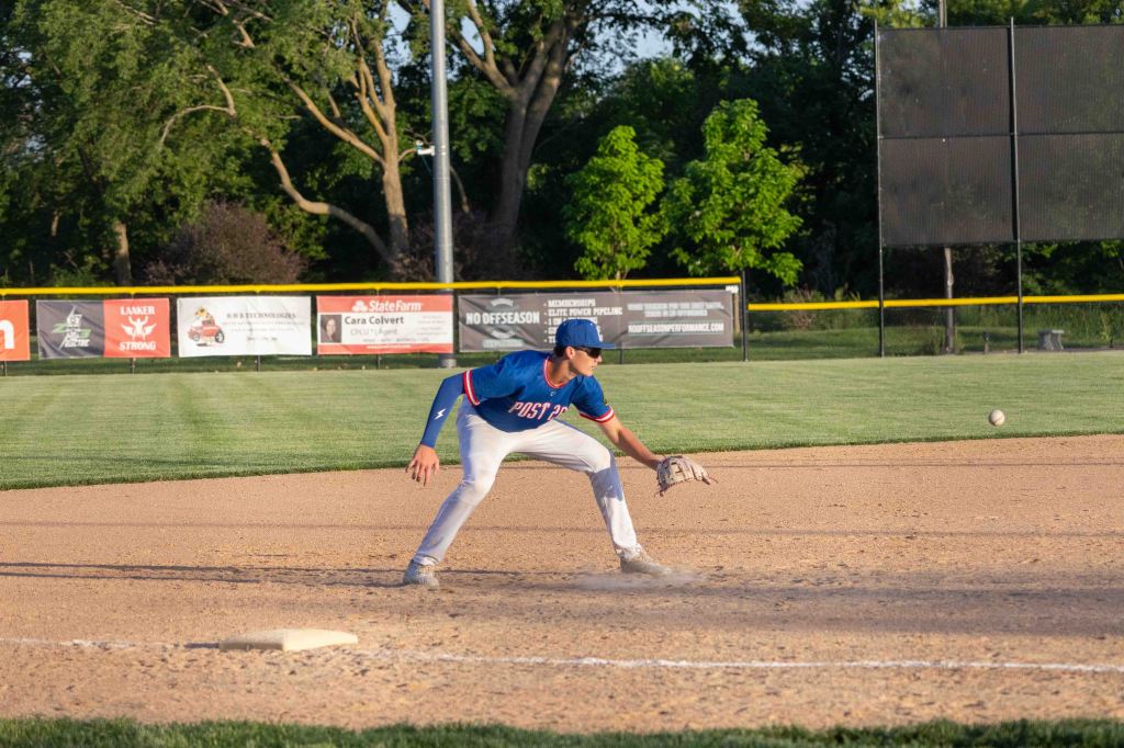 A young baseball player in a blue jersey is crouched in an ready position on a baseball field, preparing to field a ground ball.