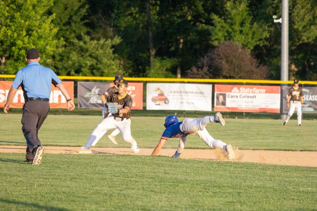 A baseball player in a blue jersey is diving towards the base, while another player in a striped black and yellow jersey prepares to catch the ball. An umpire watches the play from the foreground.