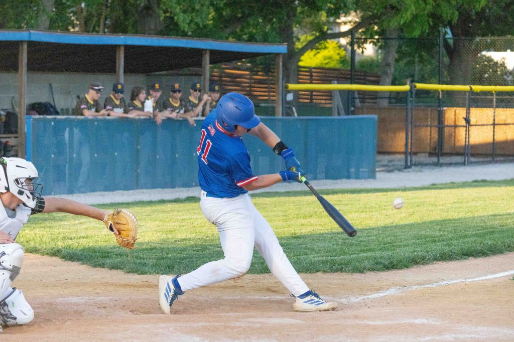 A baseball player swings at a pitch while a catcher crouches behind him, with teammates visible in the background.