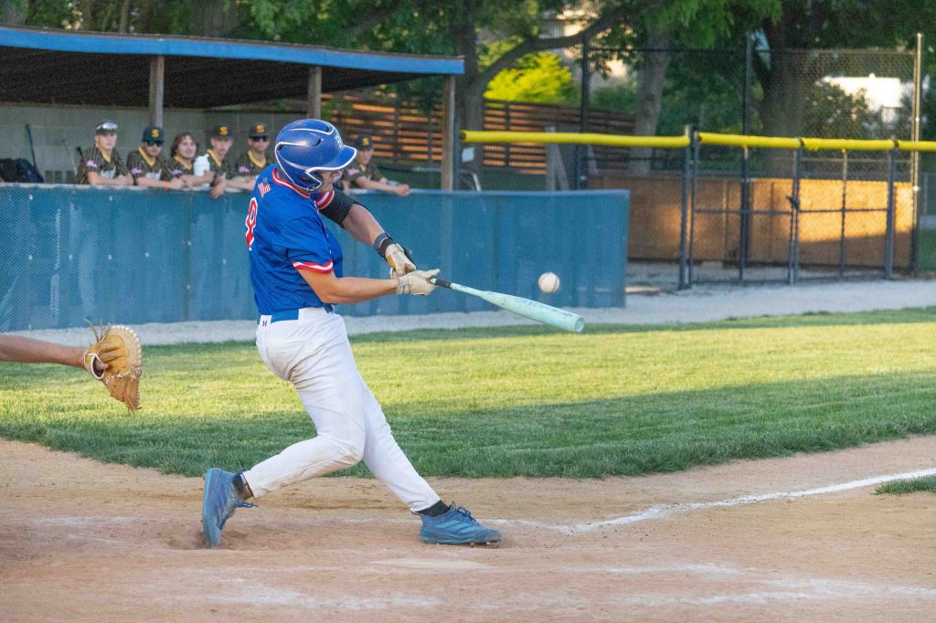 A baseball player in a blue uniform swings at a pitch as a team watches from the dugout in the background.
