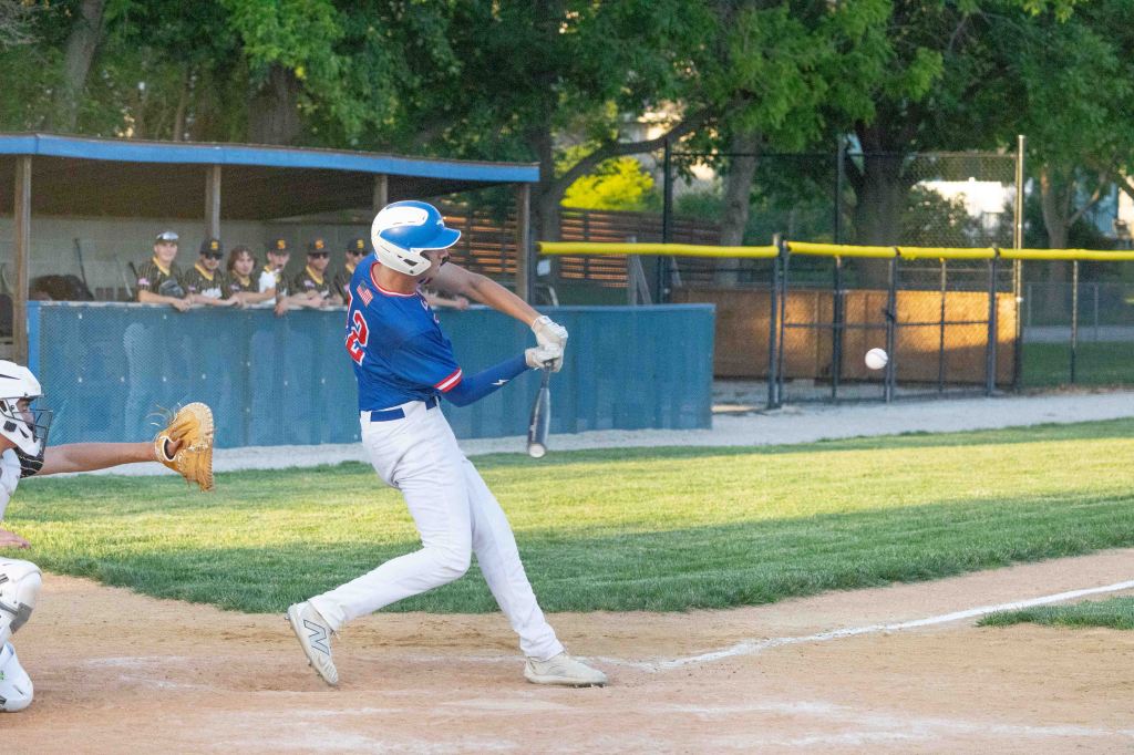 A baseball player in a blue jersey swings at a pitch, making contact with the ball, while a catcher crouches nearby. In the background, a group of players watches from the dugout.