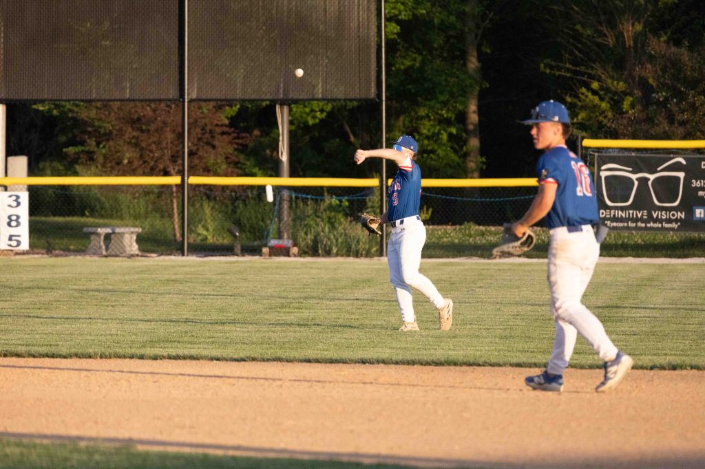 A baseball player in a blue jersey throws the ball while another player watches nearby on a sunny field.