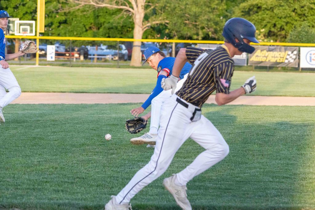 A baseball player in a blue uniform lunges for a ground ball on the field, while another player in a striped uniform runs towards first base.