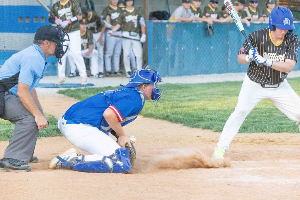 A baseball game scene featuring a batter preparing to swing, while a catcher crouches behind home plate. An umpire stands nearby, with players in the background.