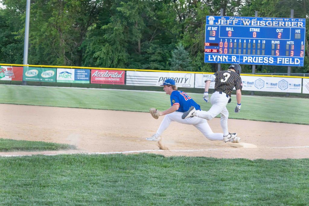A baseball player in a blue jersey stretches to catch a ball while another player in a black and gold striped jersey races towards the base, illustrating a competitive play at Lynn R. Pruess Field.