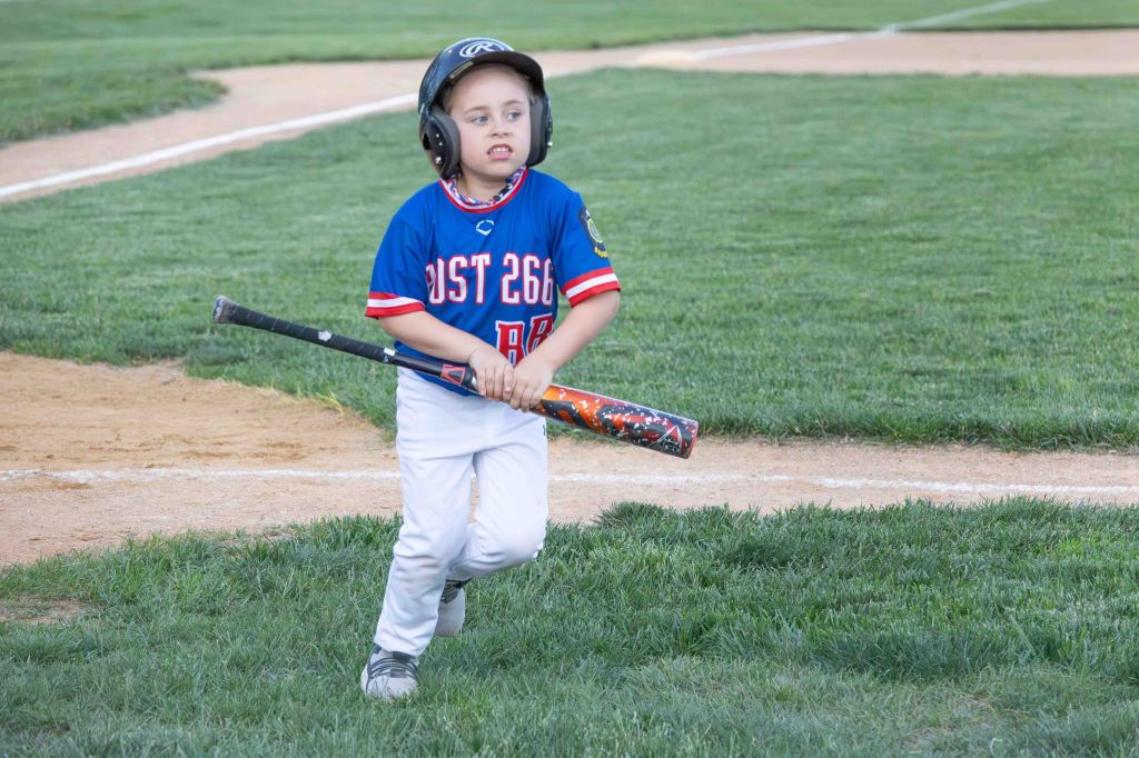 Young child in a baseball uniform holding a bat while walking on a grassy field.