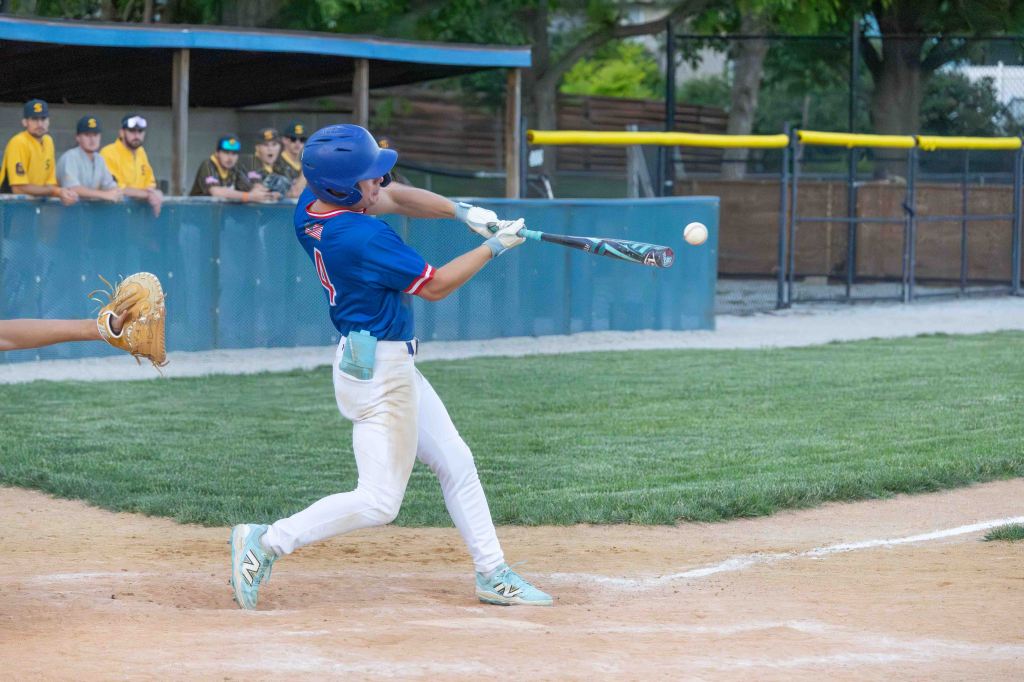A young baseball player swings a bat at a pitched ball during a game, with teammates in the background watching.