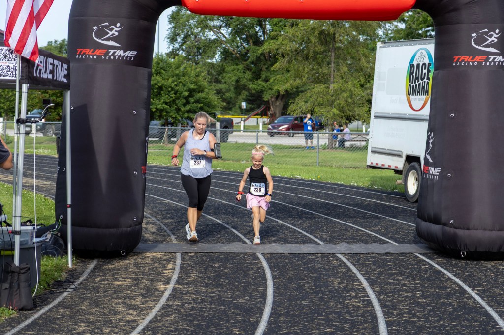A woman and a young girl running through an inflatable arch at a race finish line, with spectators in the background.
