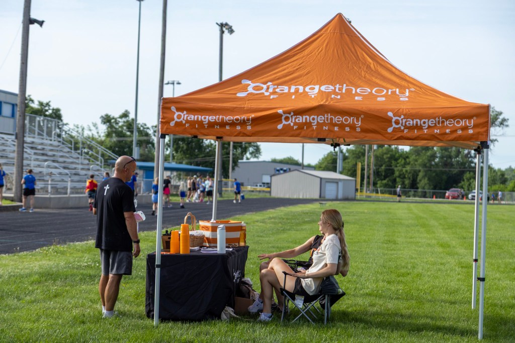 An outdoor fitness event featuring an orange tent for Orangetheory Fitness, with staff and participants engaging in activities in the background.