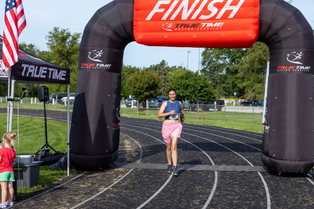 A female runner crossing the finish line under an inflatable archway with 'FINISH' written on it, at a racetrack. A small child watches nearby, and an American flag is visible in the background.