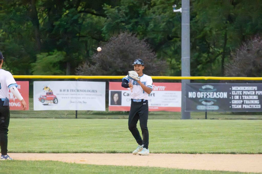 A young baseball player smiling and catching a baseball during a game on a field, with a blurred background featuring advertisements and trees.