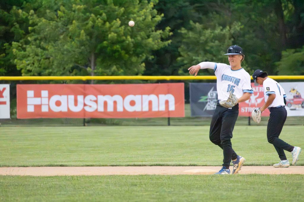 A baseball player in a white jersey, numbered 15, throws a ball while another player stands nearby on the field, with a Hausmann banner in the background.