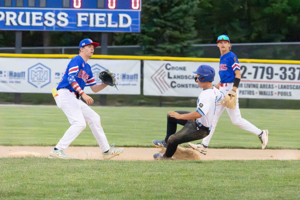 A youth baseball game scene, featuring three players. One player in a blue cap is sliding into second base, while another player in a red and blue jersey prepares to catch the ball. The scoreboard in the background shows no score.
