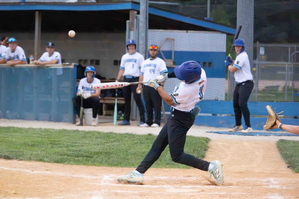 A young baseball player swings a bat to hit a pitch during a game, with teammates watching from the dugout in the background.