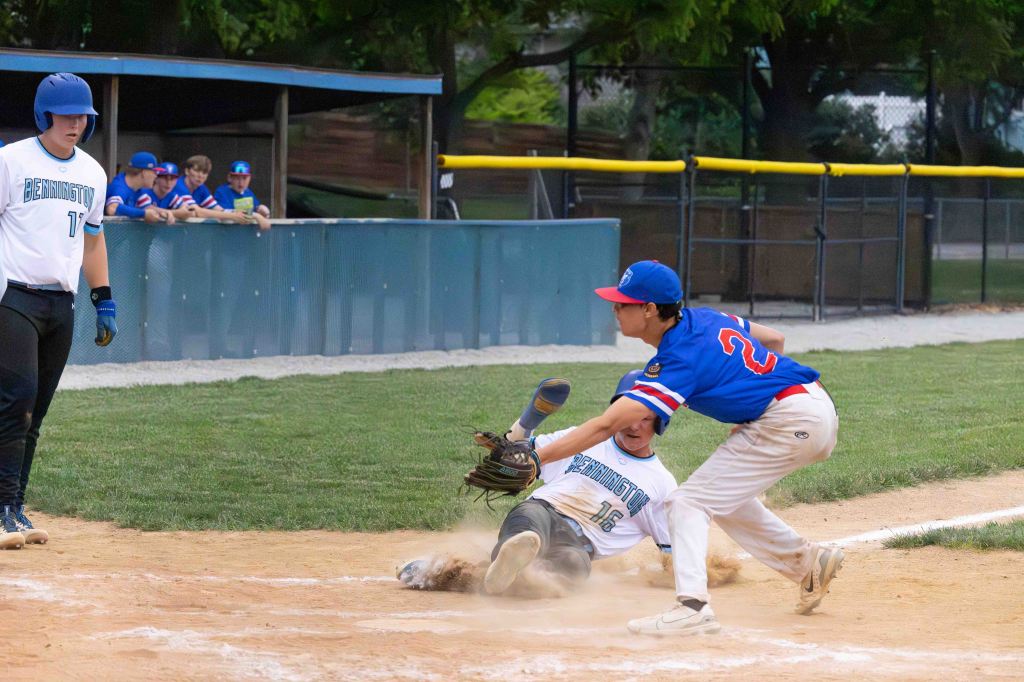 A baseball player sliding into home plate while another player attempts to tag him out. In the background, teammates watch the play from the dugout.