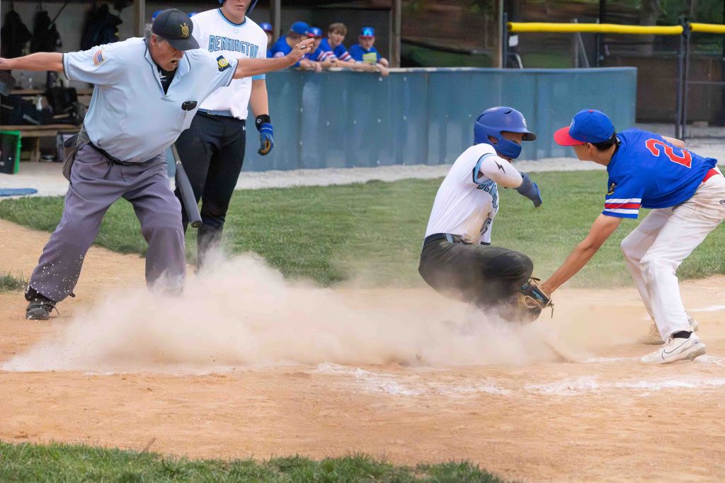 A baseball player sliding into home plate, with a catcher attempting to tag him, as the umpire raises his hands to signal a call. Dust is being kicked up from the base.