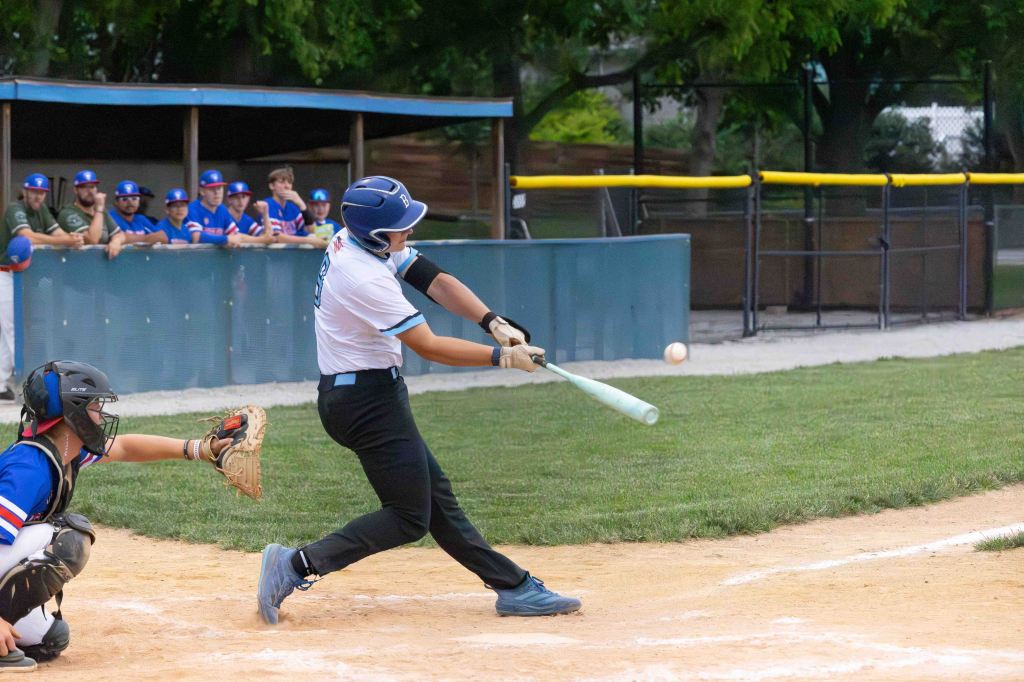 A baseball player in a white jersey swings a bat at a ball while a catcher in gear prepares to receive the pitch, with spectators in the background.