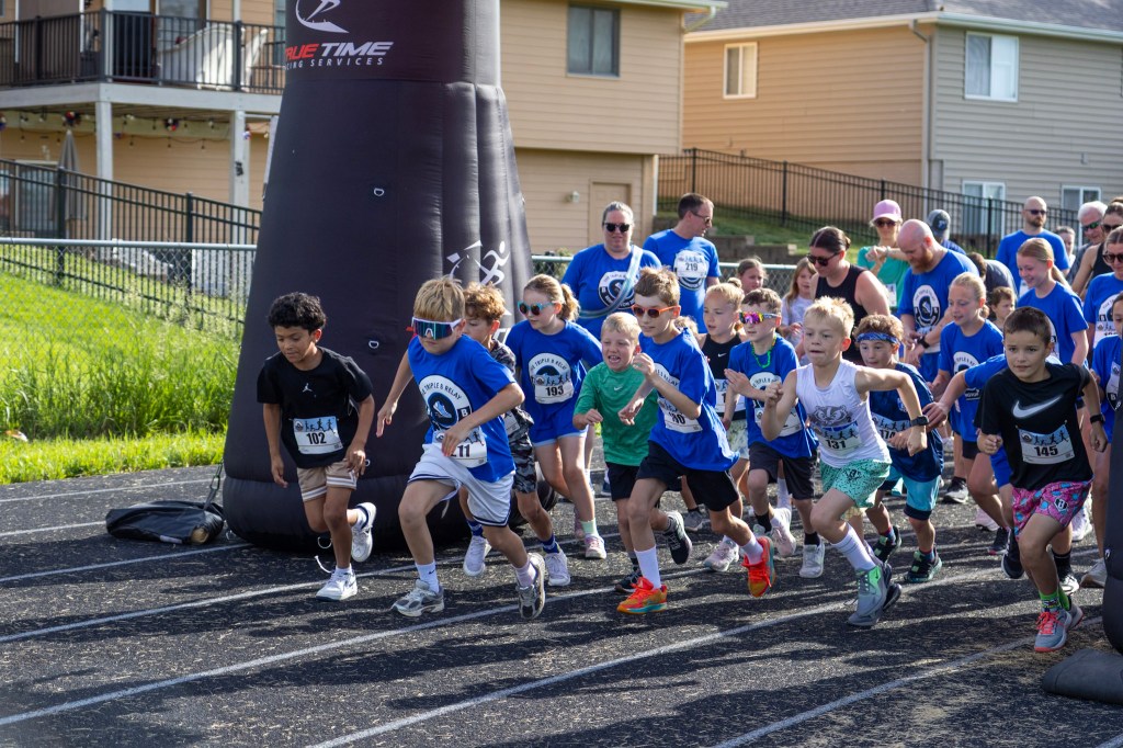 A group of children in blue shirts racing on a track at an event, with cheering spectators in the background.