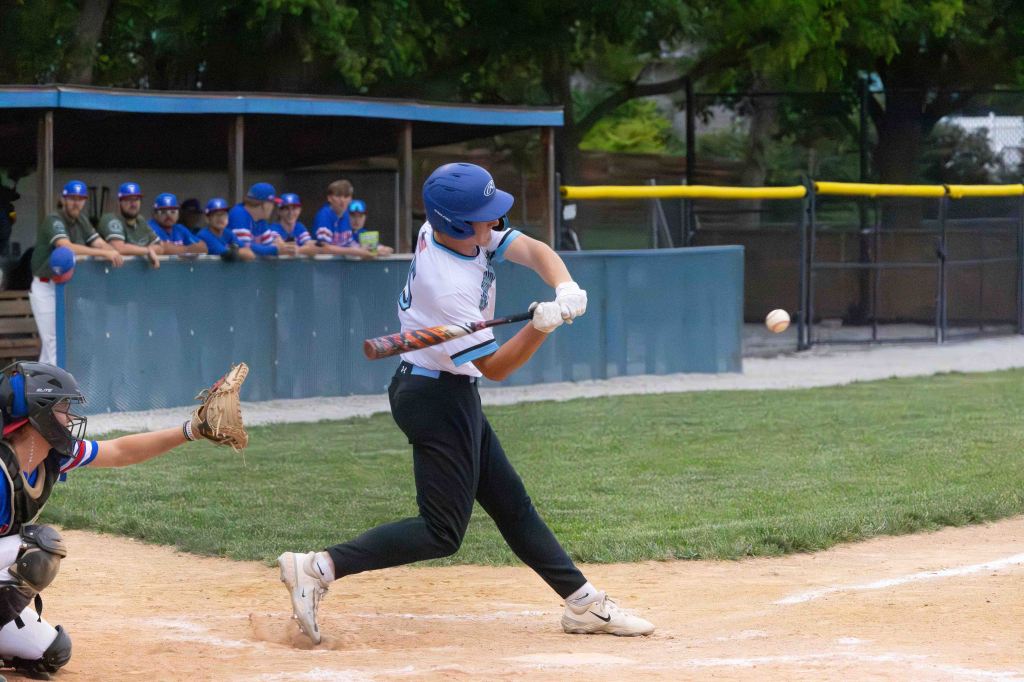 A baseball player in a blue helmet swings at a pitch, while a catcher prepares to catch the ball. Teammates are visible in the dugout in the background.