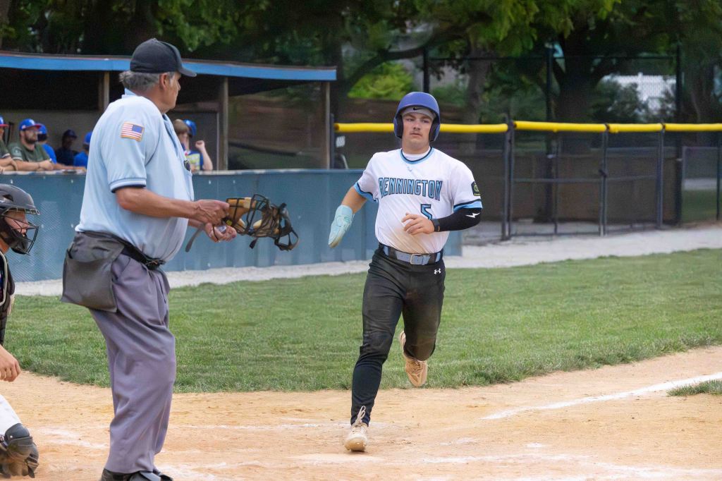 A baseball player in a white jersey with blue accents and the name 'Bennington' running towards home plate, while an umpire in blue and gray observes and a catcher in gear is positioned behind him.
