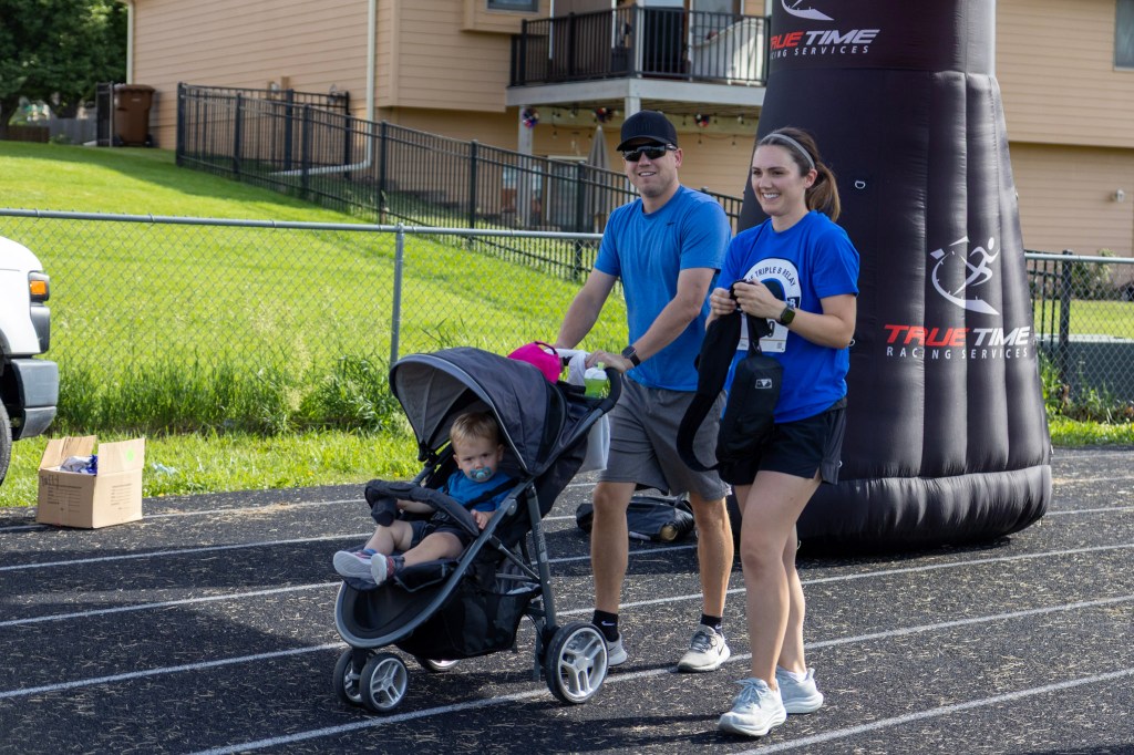 A man and a woman walk on a track, pushing a stroller with a toddler inside. The woman holds a small bag, while the man wears a cap and sunglasses. In the background, there is a large inflatable arch marked with 'True Time Racing Services' and a house with a yard.