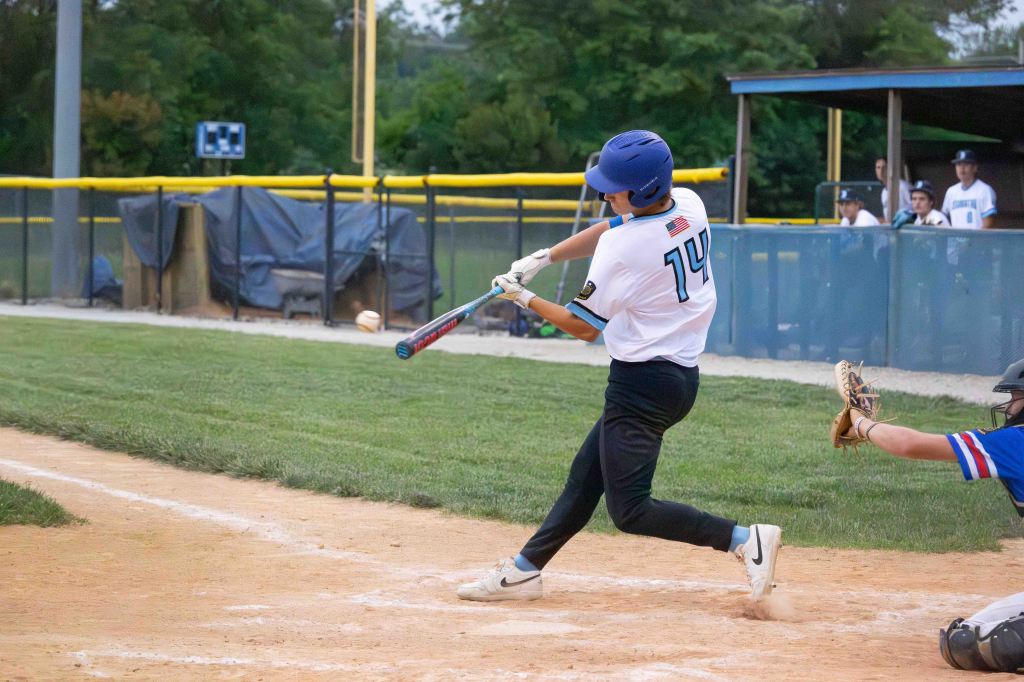 A young baseball player in a white jersey with the number 14 swings at a pitch during a game, with a catcher in gear behind him.