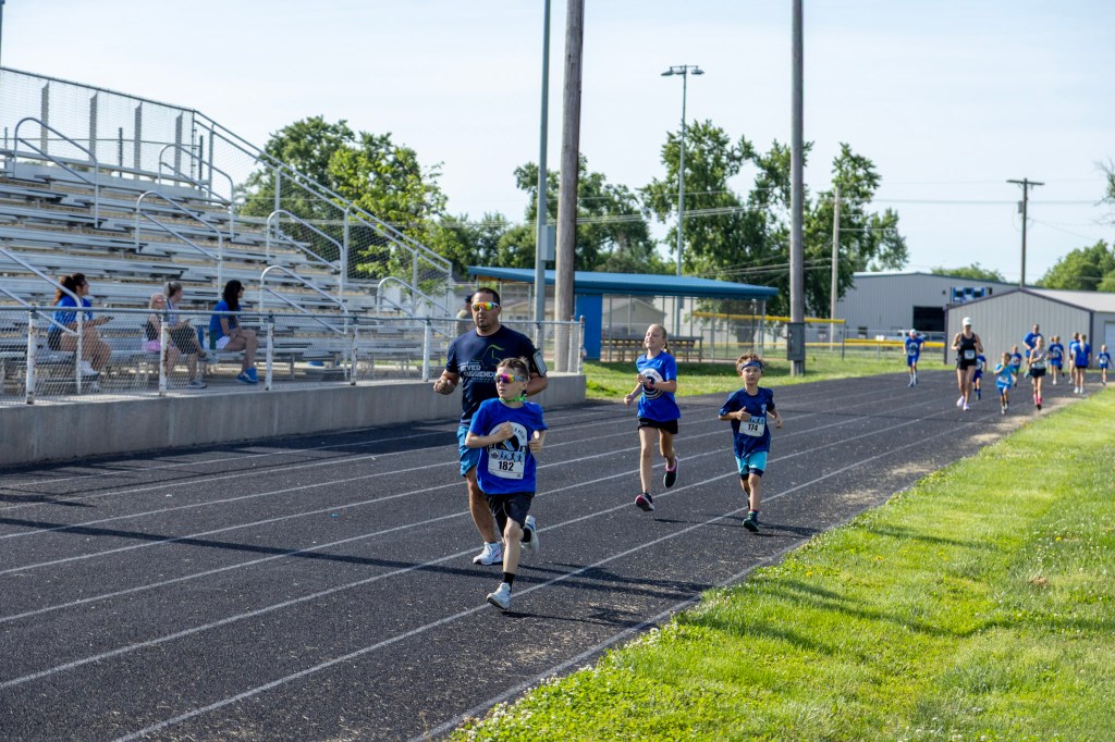 Children and an adult running on a racetrack during a community event, with spectators in the background.