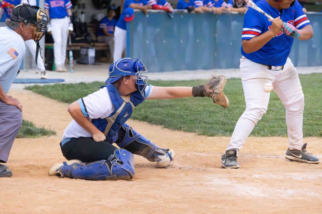 A baseball catcher crouches with one arm extended to catch a pitch, while a batter swings their bat, focusing on the game in a baseball field setting.
