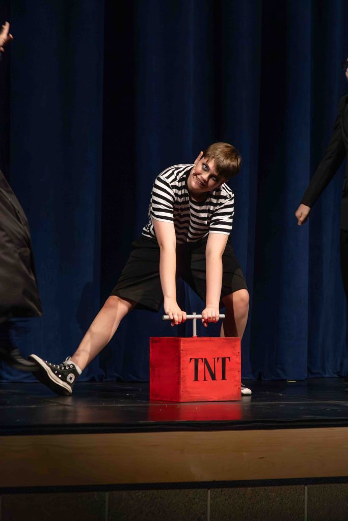 A performer in a striped shirt and black shorts is playfully pretending to lift a red TNT box on stage, surrounded by blue curtains.