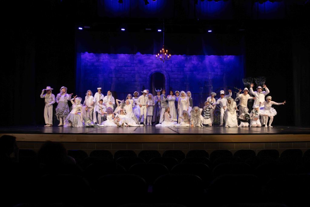 A large group of performers in white costumes poses on stage under purple lighting, with a backdrop resembling a stone wall and a chandelier above.