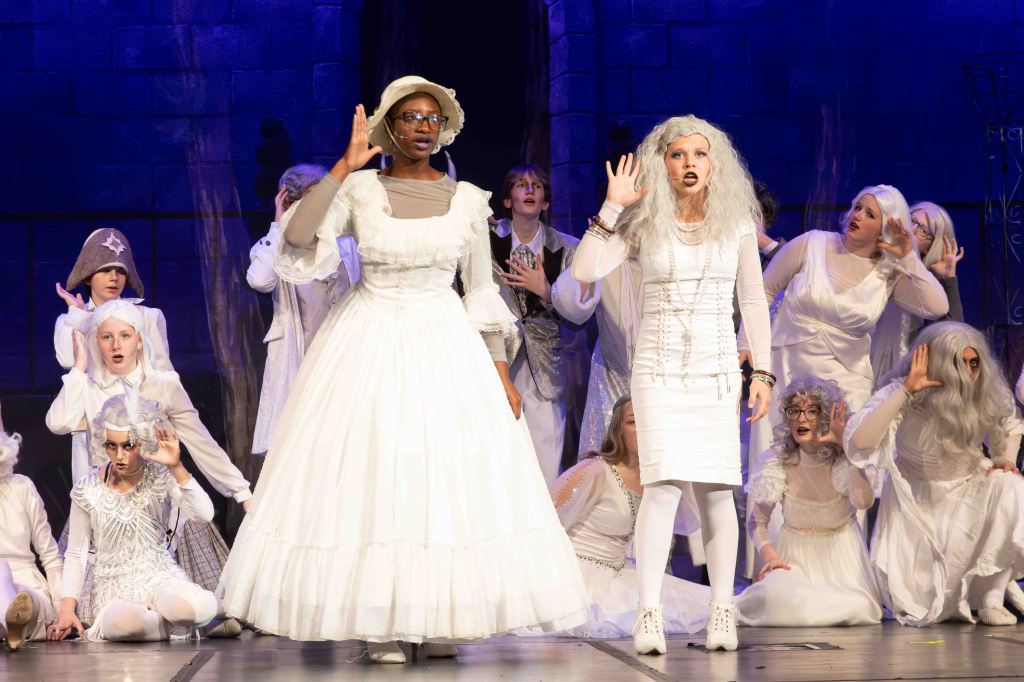 A theater performance featuring two leading characters in white costumes, surrounded by a chorus of performers in ghostly attire, all on stage with a dramatic background.