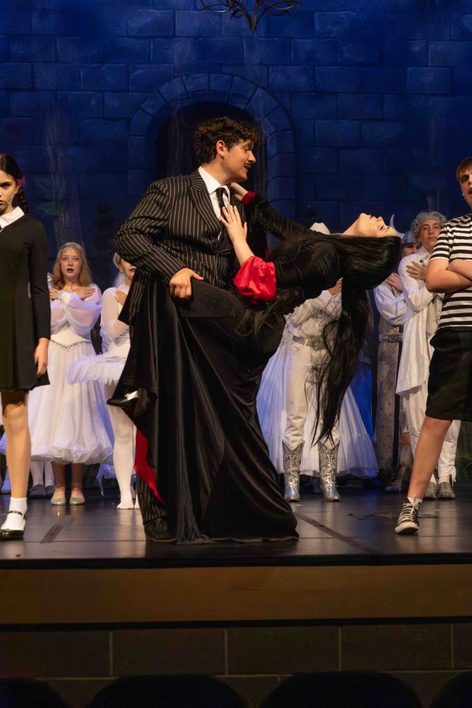A theatrical scene featuring a man in a pinstripe suit lifting a woman in a black and red dress, both performing on stage surrounded by ensemble cast members in white costumes.