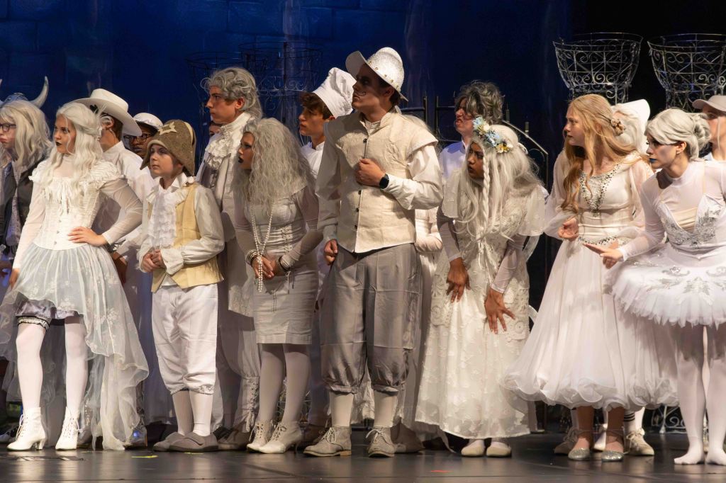 A group of performers dressed in white costumes with various accessories, standing together on stage during a theatrical production.