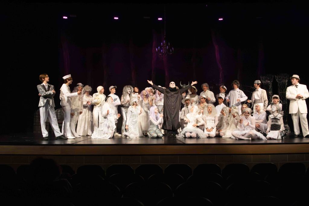A theatrical performance featuring a large cast dressed primarily in white and gray costumes, with one actor in a black outfit portraying a central character. The backdrop is dimly lit, creating a dramatic atmosphere on stage.