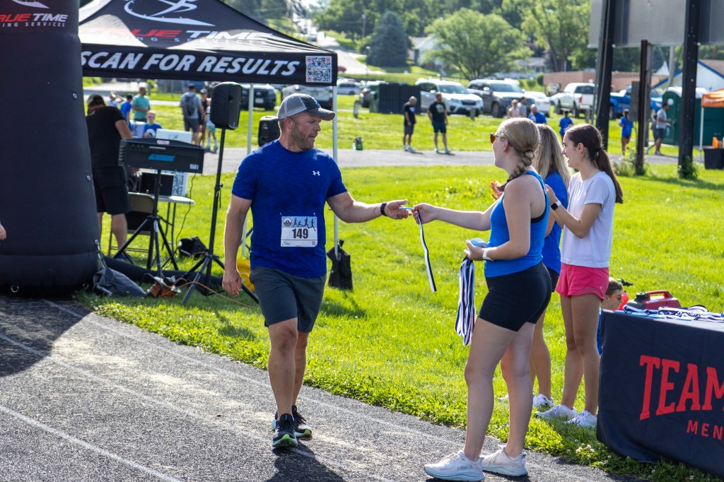 A man receiving a medal from a woman at a race event, with spectators and a registration area in the background.