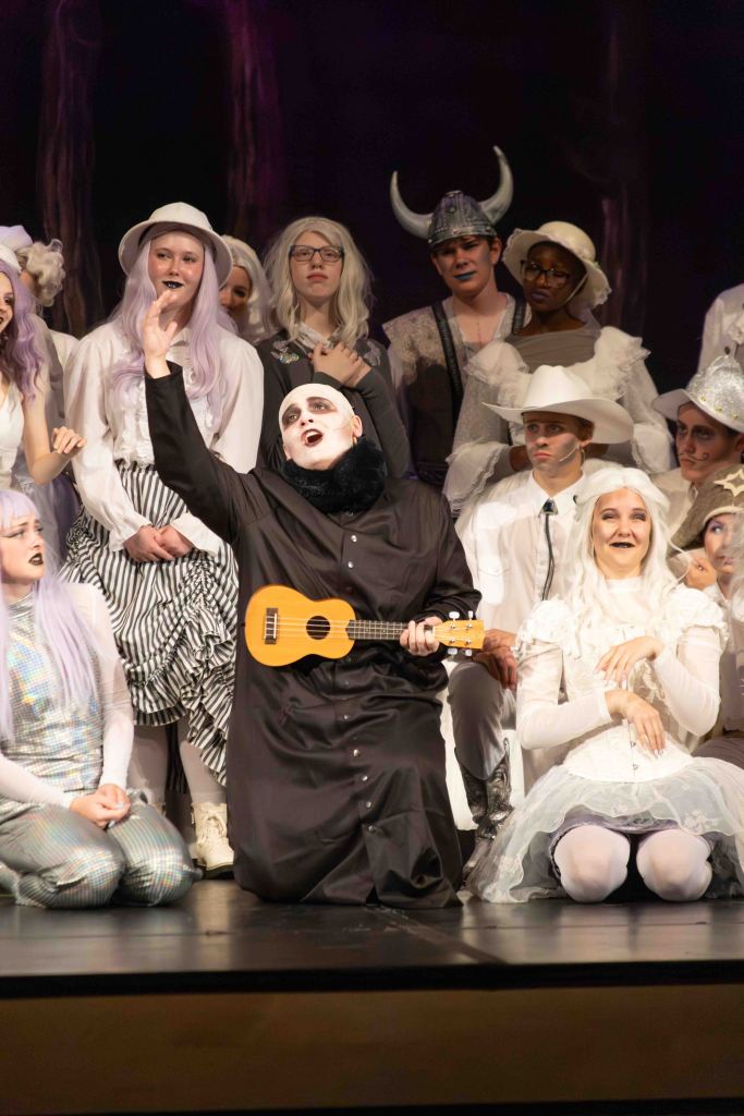 A performer in a black outfit kneels on stage holding a ukulele, surrounded by a diverse cast of actors in whimsical costumes, some with white hair and hats, during a theatrical performance.