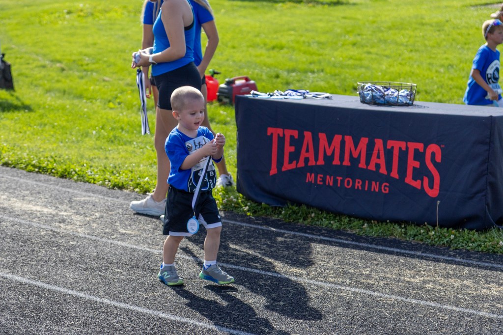 A young boy in a blue shirt and black shorts smiles while holding a medal on a running track, with a 'TEAMMATES MENTORING' banner in the background.