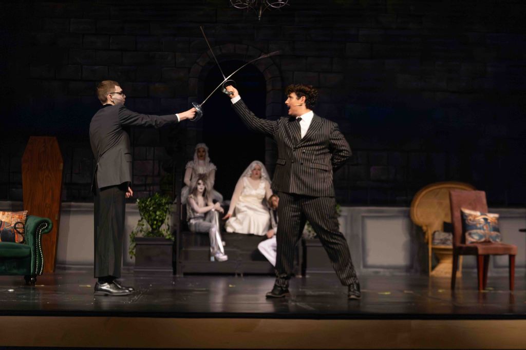 Two actors in formal attire engage in a sword duel on stage, with a dramatic backdrop and other characters seated in the background.