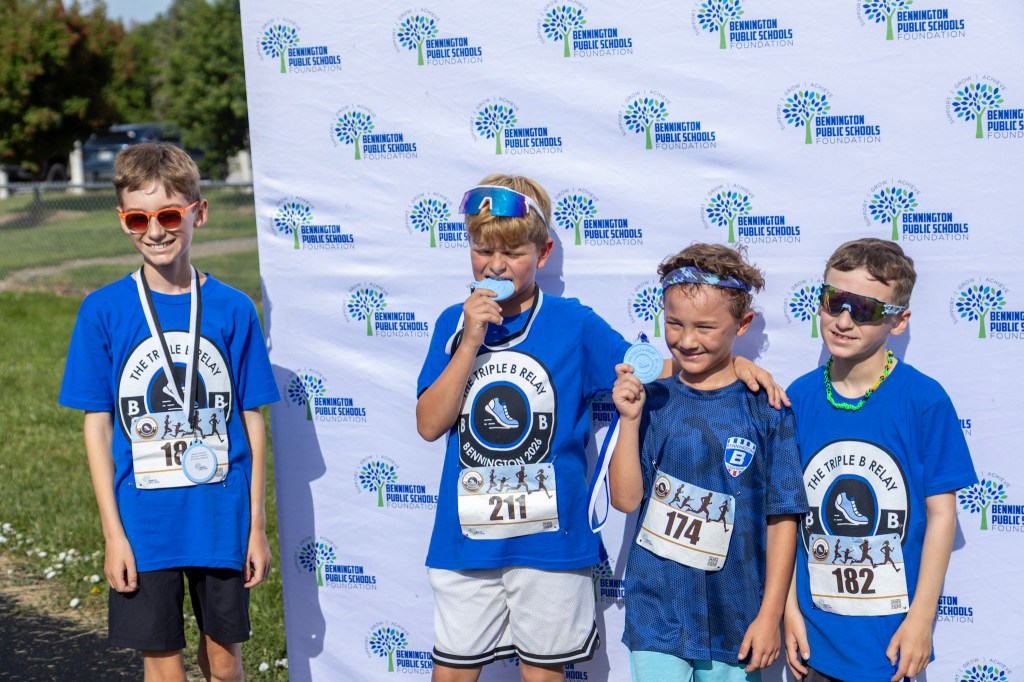 Four young runners celebrating at an event, each wearing blue shirts with race numbers, standing in front of a backdrop that reads 'Bennington Public Schools Foundation'. Three of them hold medals.