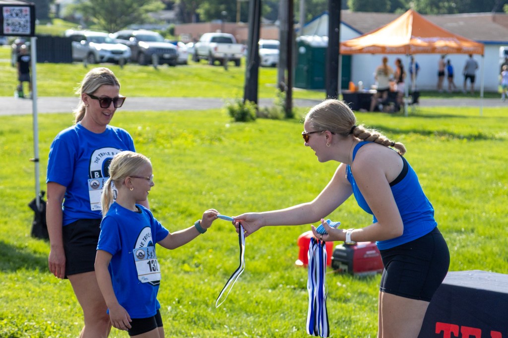 A young girl receives a medal from an older girl during an award ceremony, with two women in blue shirts watching in the background.