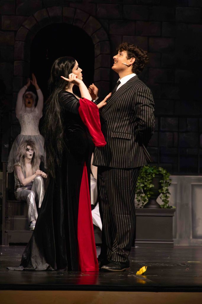 A theatrical scene featuring a woman in a black and red dress interacting with a man in a striped suit. They share a moment of playful expression, while ghostly figures and a stone archway form the background.