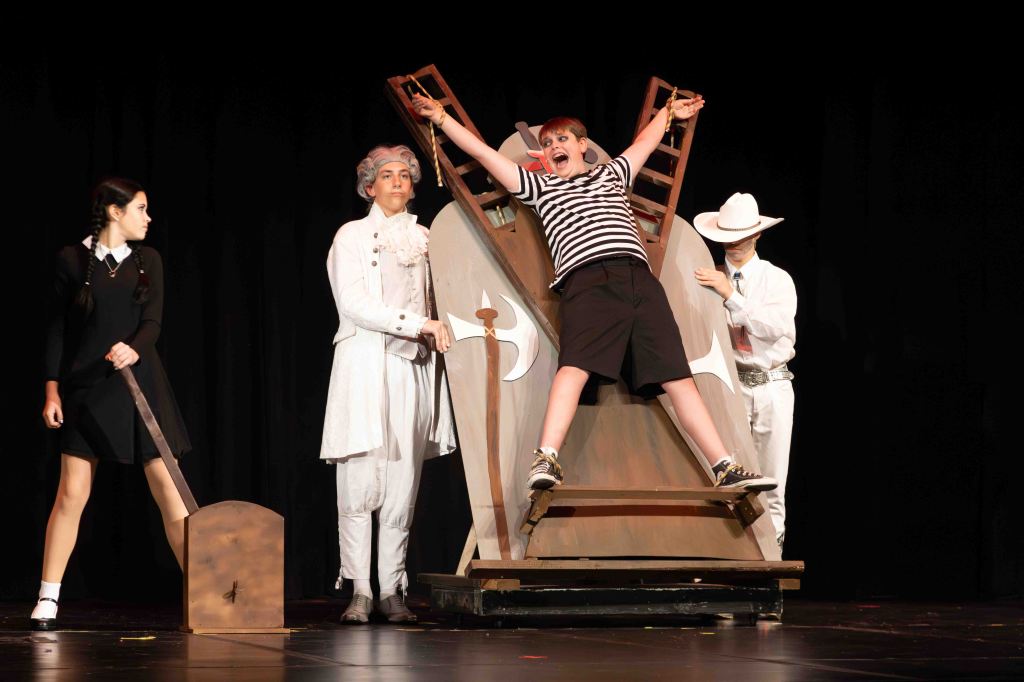 A scene from a theatrical performance featuring four characters: a boy in striped clothing being lifted in an exaggerated manner by a prop resembling a guillotine, flanked by two actors in historical costumes and a girl in a black dress.