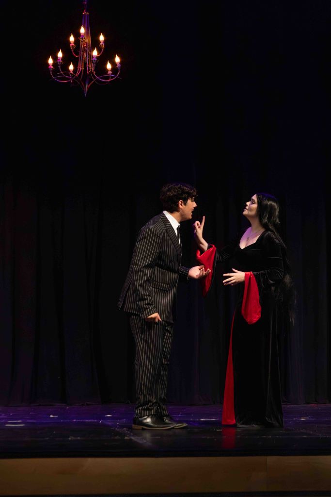 A scene from a theatrical performance featuring a man in a striped suit and a woman in a black and red gown, engaged in a dramatic conversation under a chandelier.