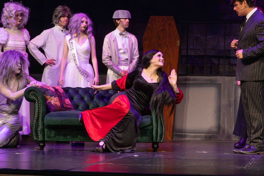 A theatrical performance featuring several characters in costumes, including a woman in a black dress with red accents lounging on a green couch, while other performers in various outfits stand nearby.