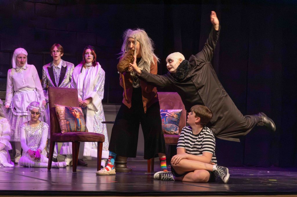 A theatrical scene featuring a group of actors in dramatic costumes, with one actor in a dark robe being playfully restrained by another actor in a vibrant outfit. Several other actors observe from the background, and a child sits on the floor, watching the performance.