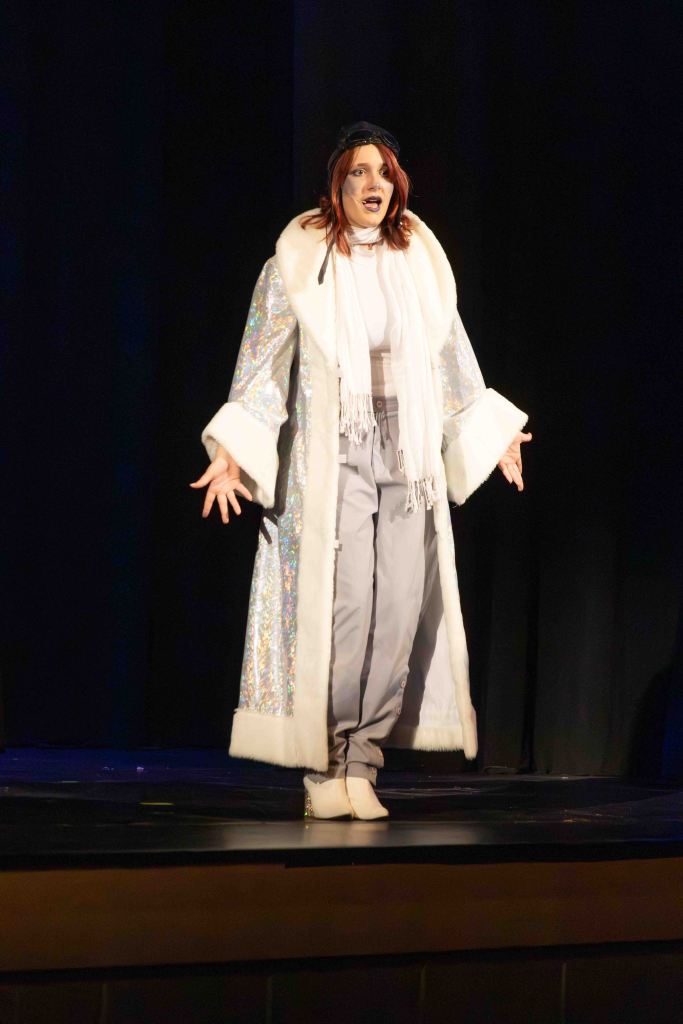 A performer in a glittering long coat with fur trim, gray pants, and white boots, passionately engaging with the audience on stage.