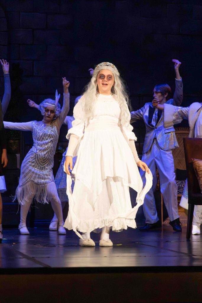A performer in a white dress and long wig stands on stage, surrounded by other dancers in theatrical costumes, celebrating a lively moment in a play.